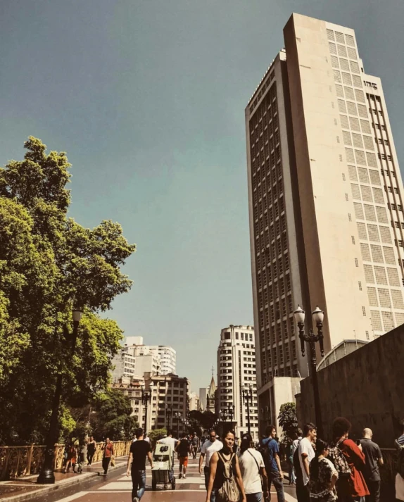 a group of people walking down a street next to tall buildings, an album cover, by Alejandro Obregón, unsplash, brutalism, with a tall tree, chile, visible from afar!!, instagram picture