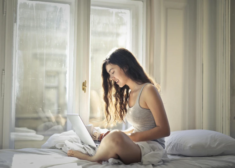 a woman sitting on a bed using a laptop computer, pexels contest winner, happening, wearing a camisole and shorts, window light, wearing pajamas, handsome girl