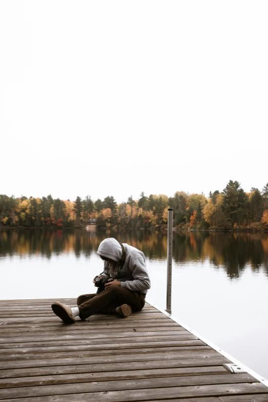 a person sitting on a dock next to a body of water, by Jaakko Mattila, during autumn, camera photo, gray men, hooded