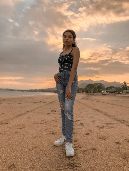 a woman standing on top of a sandy beach, by Robbie Trevino, wearing jeans, ((sunset)), background image, full body cute young lady