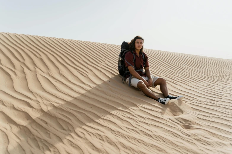 a man sitting on top of a sand dune, trending on pexels, with a backpack, tanned ameera al taweel, male teenager, australian