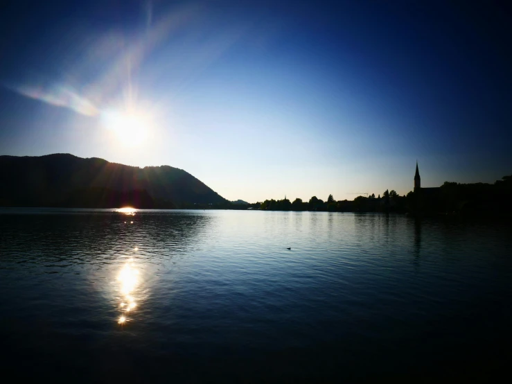 the sun is setting over a body of water, by Niko Henrichon, pexels contest winner, lakeside mountains, clear sunny day, blue sky, blurred