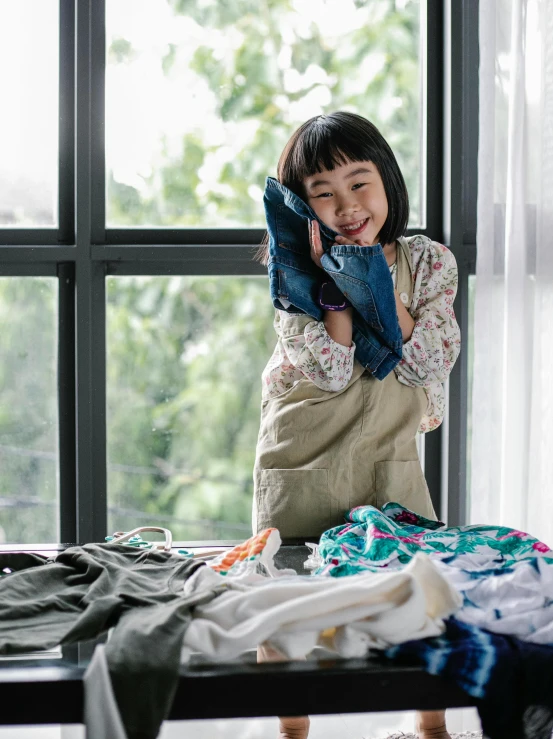 a little girl standing on top of a bed next to a pile of clothes, by Jang Seung-eop, pexels contest winner, girl making a phone call, wearing an apron, inspect in inventory image, laundry hanging