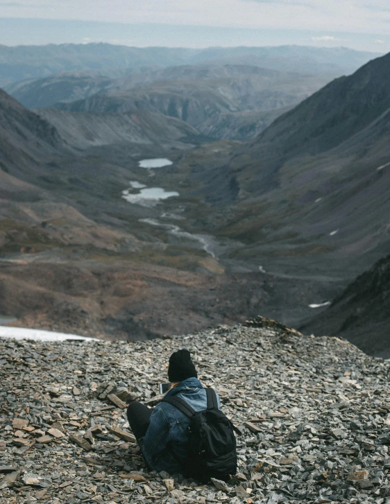 a person sitting on top of a rocky hill, looking down at the valley, gravel and scree ground, with a backpack, /r/earthporn