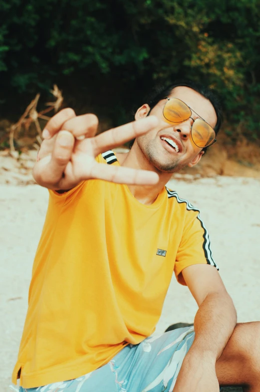 a man sitting on top of a skateboard on a beach, wearing gold glasses, holding his hands up to his face, wearing a modern yellow tshirt, mohamed chahin style