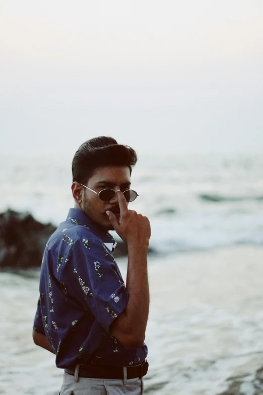 a man standing on top of a beach next to the ocean, an album cover, unsplash, wear ray - ban glass, indian, male teenager, profile image