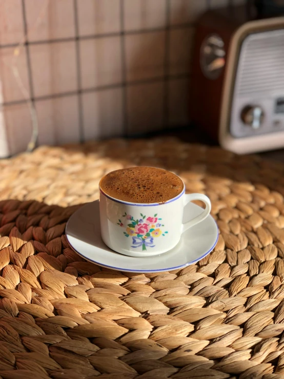 a cup of coffee sitting on top of a wicker table, by Lucia Peka, brazilian, promo image, fan favorite, stroopwaffel