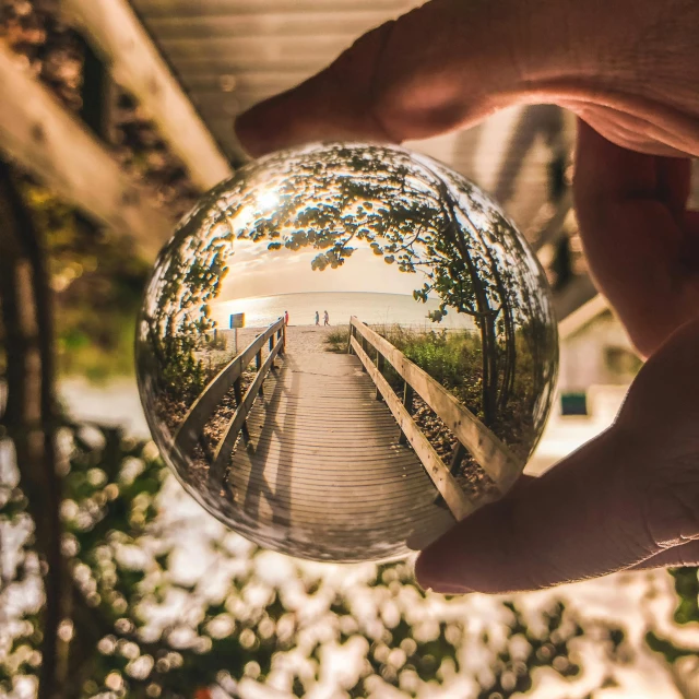 a person holding a glass ball with a bridge in the background, by Jakob Gauermann, unsplash contest winner, photorealism, fisheye!!! lens, beach surreal photography, tree of life inside the ball, instagram post