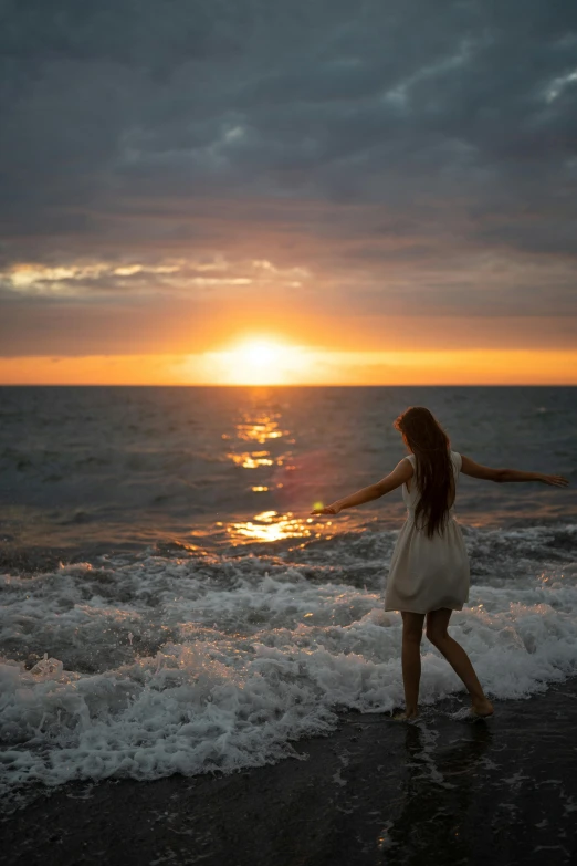 a woman standing on top of a beach next to the ocean, during a sunset, whirling, kailee mandel, in the ocean