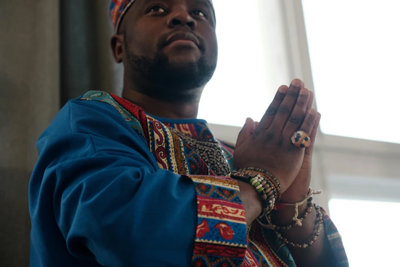 a man is praying in front of a window, an album cover, inspired by Ras Akyem, pexels contest winner, hurufiyya, wearing an ornate outfit, wakanda, 2 5 6 x 2 5 6 pixels, blessing hands