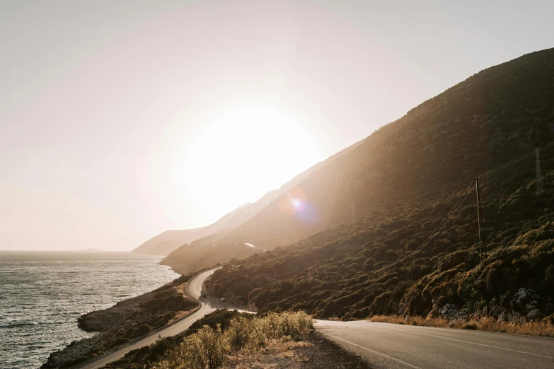 a motorcycle parked on the side of a road next to the ocean, by Lee Loughridge, unsplash contest winner, sun rises between two mountains, hollister ranch, hazy sun and mystical, landslide road