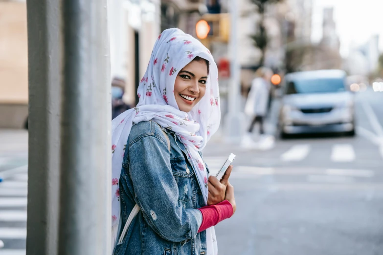 a woman standing on the side of a street holding a cell phone, a photo, hurufiyya, patterned scarf, gen z, welcoming grin, modest