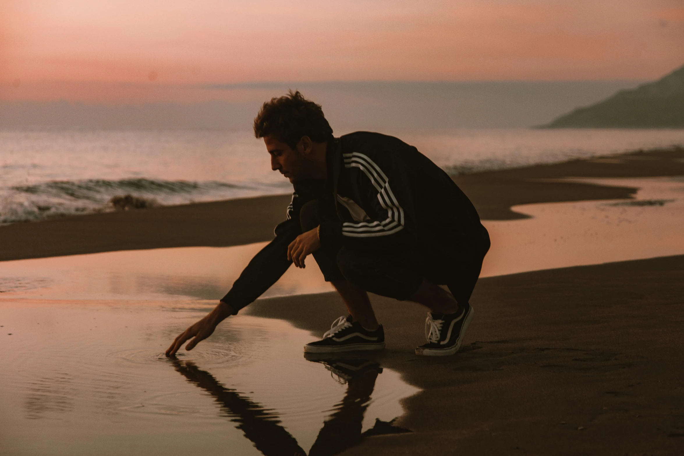 a man kneeling on top of a beach next to the ocean, pexels contest winner, chappie in an adidas track suit, high quality reflections, flume, soft glow