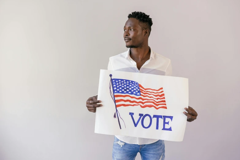 a man holding a sign that says vote, a photo, trending on pexels, black arts movement, stars and stripes, handsome man, young thug, on a pale background