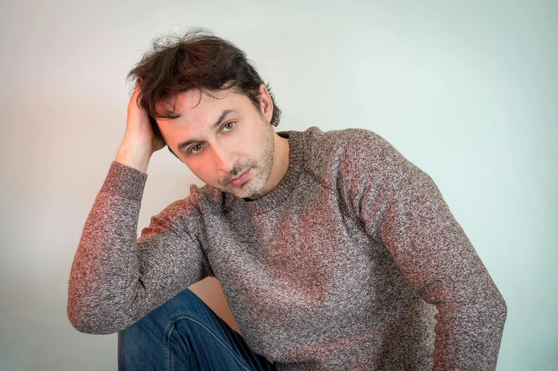 a man sitting on the floor with his hand on his head, inspired by Joaquín Torres García, pexels contest winner, photorealism, he is wearing a brown sweater, avatar image, in front of white back drop, light stubble with red shirt