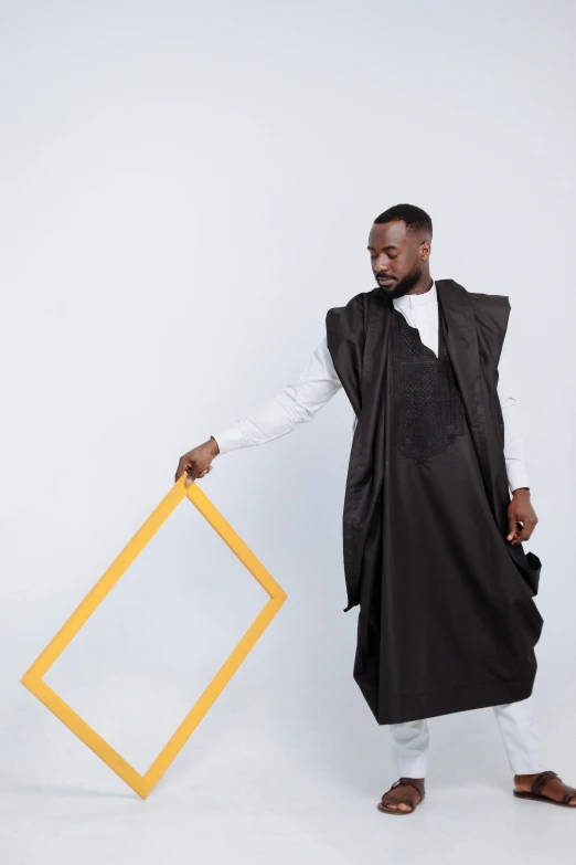 a man standing in front of a white wall holding a yellow frame, an album cover, inspired by Ras Akyem, pexels contest winner, wearing black robes, model is wearing techtical vest, doing an elegant pose over you, square