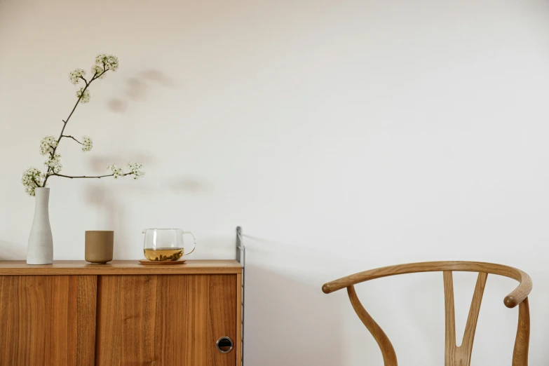 a wooden cabinet sitting next to a wooden chair, inspired by Constantin Hansen, trending on unsplash, minimalism, ikebana, background image, table in front with a cup, white background wall