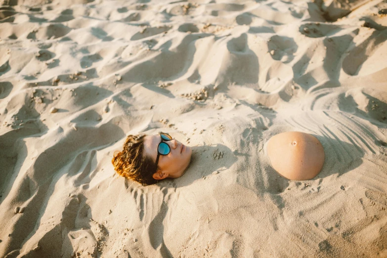 a young boy laying on top of a sandy beach, by Elsa Bleda, unsplash contest winner, surrealism, big heads, large breasts, with sunglasses, life size