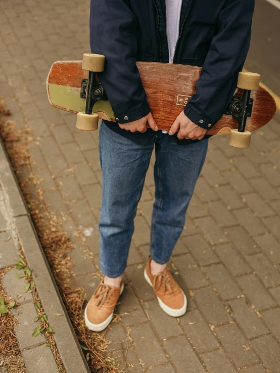 a man standing on a sidewalk holding a skateboard, a picture, by Niko Henrichon, natural wood top, ( ( ( wearing jeans ) ) ), brown ) ), handcrafted