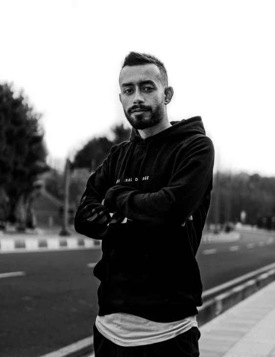 a man standing on a sidewalk with his arms crossed, a black and white photo, by Ismail Acar, wearing a black hoodie, discord profile picture, thiago alcantara, profile image