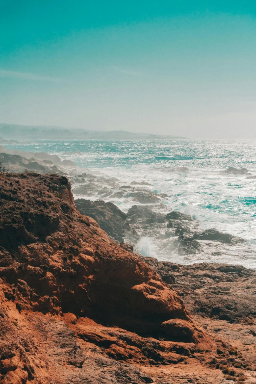 a man standing on top of a rocky beach next to the ocean, red dusty soil, epic coves crashing waves plants, slide show