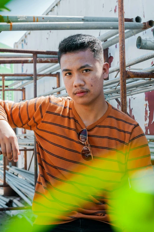 a man in an orange shirt sitting on a scaffolding, a portrait, pexels contest winner, sumatraism, he is about 20 years old | short, promo photo, caramel. rugged, patterned