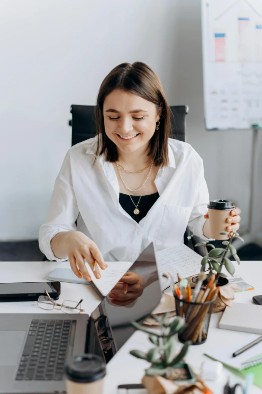 a woman sitting at a desk with a laptop and papers, welcoming grin, curated collections, wearing a white blouse, avatar image