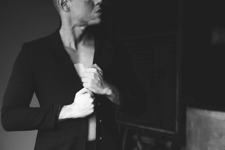 a black and white photo of a man adjusting his tie, by Emma Andijewska, wearing a black catsuit, shaved head, queer woman, jacket over bare torso