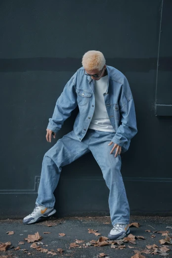 a man leaning against a wall with his hands on his hips, an album cover, unsplash, photorealism, pale blue outfit, baggy jeans, die antwoord style wear, denim jacket