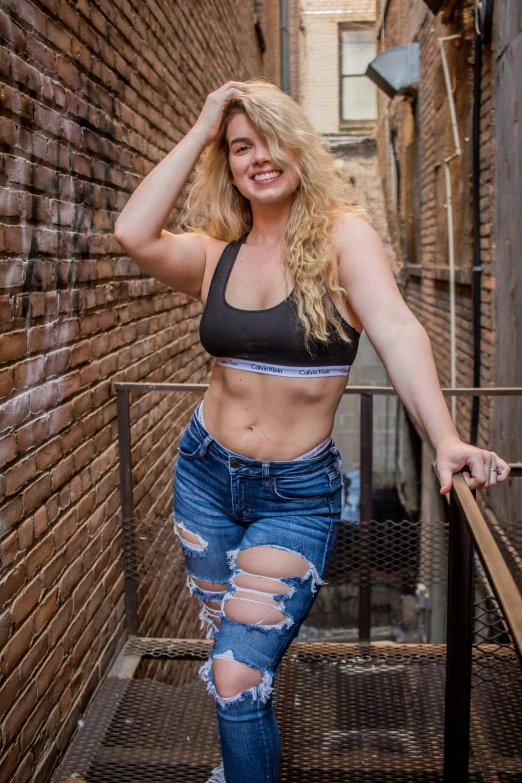 a woman posing for a picture in front of a brick wall, ripped clothing, curly blonde hair, large)}], posing in bra