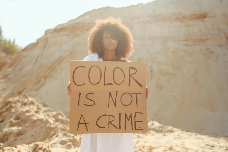 a woman holding a sign that says color is not a crime, trending on pexels, sand color, essence, crimes, ashteroth