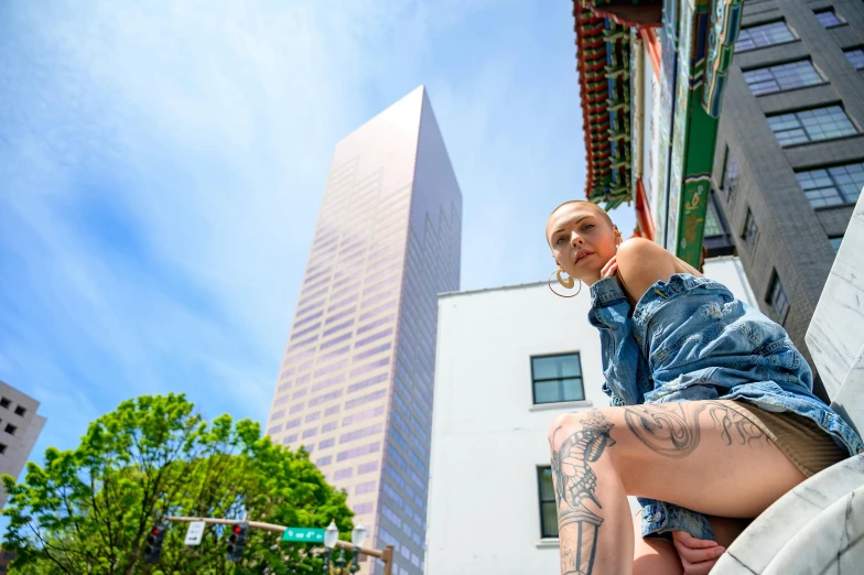 a woman sitting on a ledge in front of a tall building, inspired by David LaChapelle, pexels contest winner, hyperrealism, tattooed, clear blue skies, looking towards camera, tattoo parlor photo