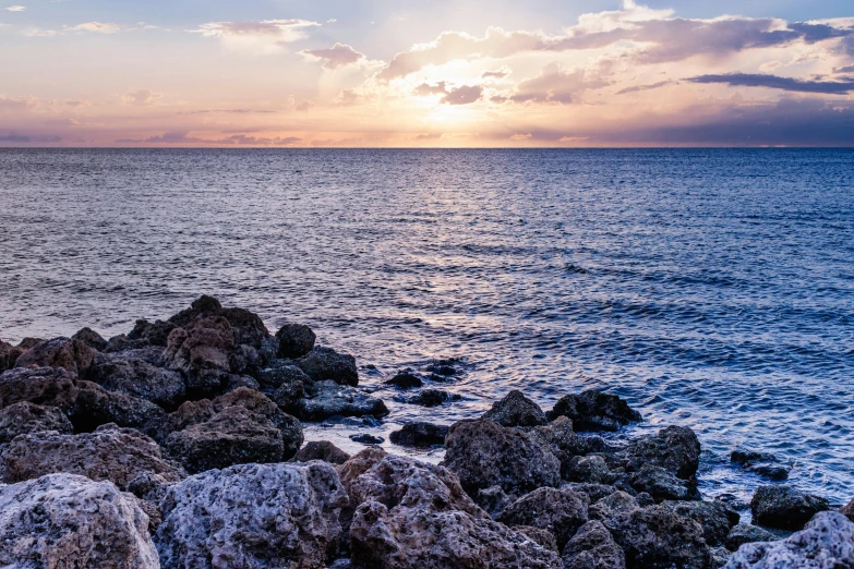 a sunset over the ocean with rocks in the foreground, by Pamela Drew, unsplash, cyprus, blue sea, jamaica, digitally enhanced