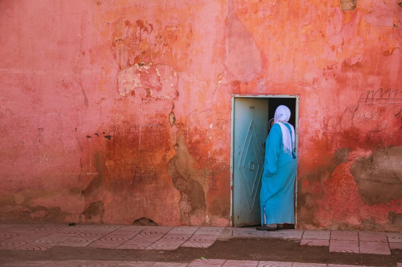 a woman in a blue dress standing in a doorway, an album cover, inspired by Steve McCurry, unsplash contest winner, moroccan city, pink and orange, burka, solid colours