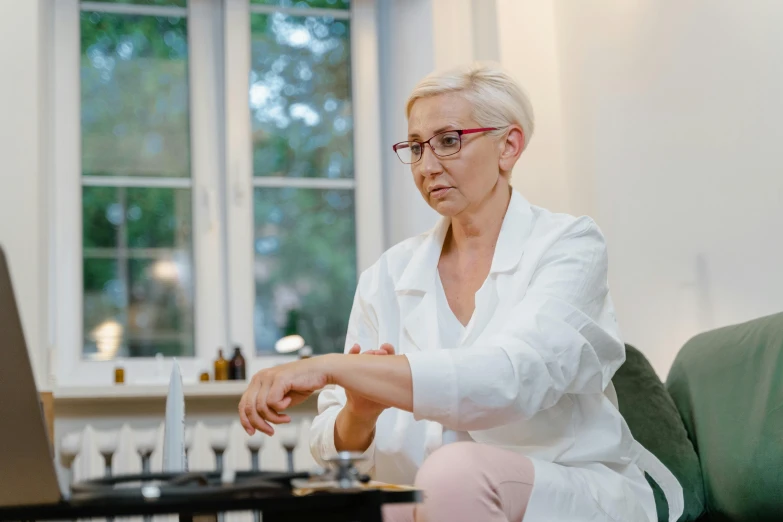 a woman sitting on a couch using a laptop computer, a hyperrealistic painting, by Emma Andijewska, unsplash, hyperrealism, wearing lab coat and glasses, partially cupping her hands, white haired lady, acupuncture treatment