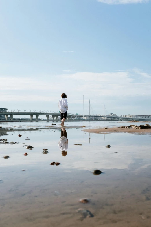 a man standing on a beach next to a body of water, by Elizabeth Durack, minimalism, bridges, teenage girl, coastal, reflect