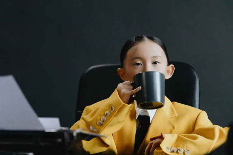 a little boy sitting at a desk drinking from a cup, inspired by Cao Buxing, pexels contest winner, girl in suit, yellow and black, young asian girl, avatar image