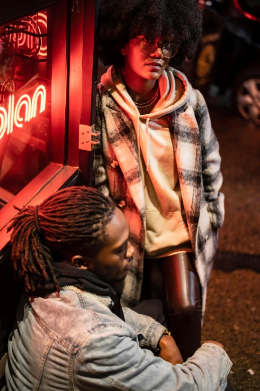 a man sitting on the ground next to a woman, by Raphaël Collin, trending on unsplash, red neon lights, model wears a puffer jacket, nubian, in london