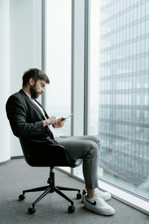 a man sitting in a chair looking at a tablet, pexels contest winner, business outfit, big windows, plain background, corporate phone app icon