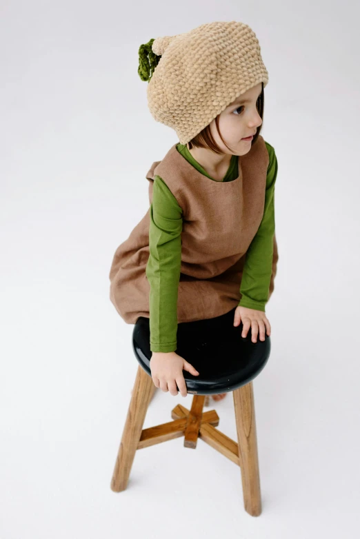 a little girl sitting on top of a wooden stool, inspired by Elsa Beskow, unsplash, renaissance, sleeveless turtleneck, green and brown clothes, beanie, thumbnail