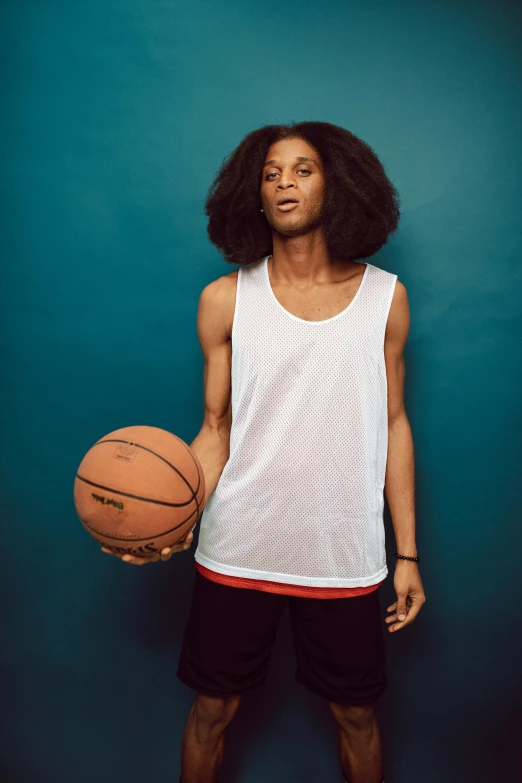 a man standing in front of a blue wall holding a basketball, by Nathalie Rattner, featured on dribble, long afro hair, sleeveless tops, promotional image, teenage boy