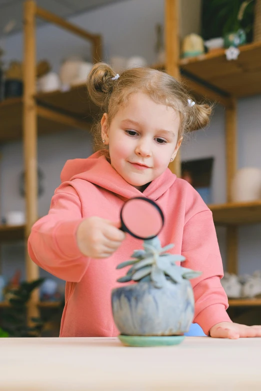 a little girl sitting at a table holding a magnifying glass, wearing a pastel pink hoodie, next to a plant, 2019 trending photo, looking at the treasure box