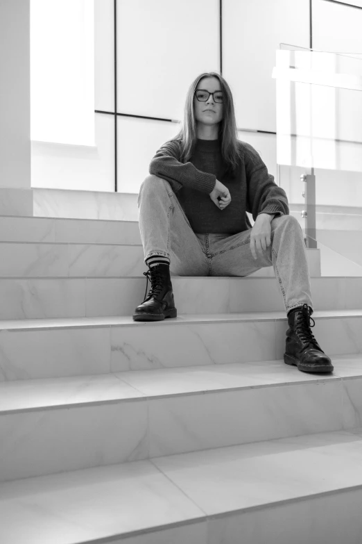 a woman sitting on the steps of a building, a black and white photo, unsplash, hyperrealism, with square glasses, androgynous male, young with long hair, standing in corner of room