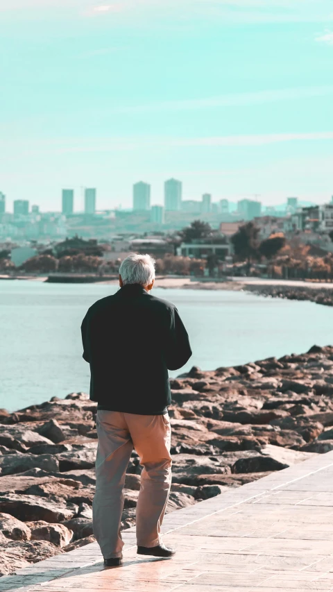 a man standing on top of a pier next to a body of water, looking at the city, grandfatherly, unsplash transparent, standing on rocky ground