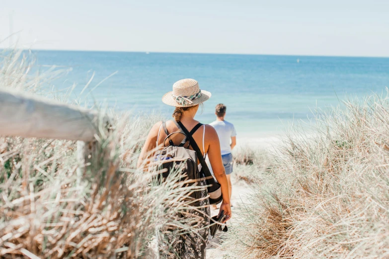 a woman walking down a path next to the ocean, by Terese Nielsen, unsplash, on a hot australian day, hunting, over his shoulder, the emerald coast