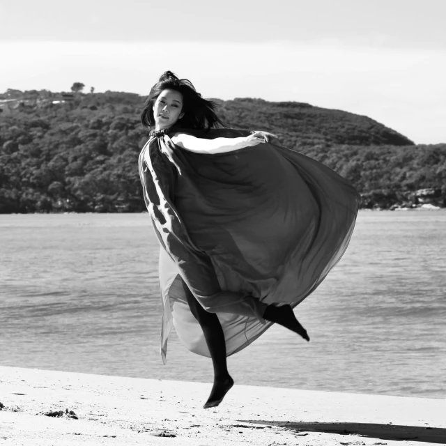 a black and white photo of a woman on the beach, figuration libre, long flowing cape and cowl, flying in the air, in a beachfront environment, wearing a wonderful dress