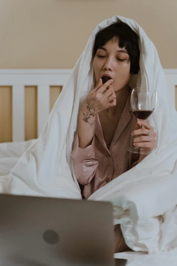 a woman sitting on a bed with a laptop and a glass of wine, cloak covering face, licking out, chill vibe, wearing simple robes