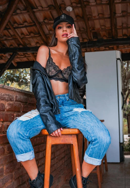 a woman sitting on top of a wooden stool, by Robbie Trevino, trending on pexels, leather jacket and denim jeans, in a black betch bra, brazilan supermodel, cold as ice! 🧊