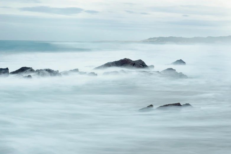 a large body of water with rocks in the foreground, by Andrew Geddes, unsplash contest winner, tonalism, pale blue fog, sea spray, medium format. soft light, motion lines