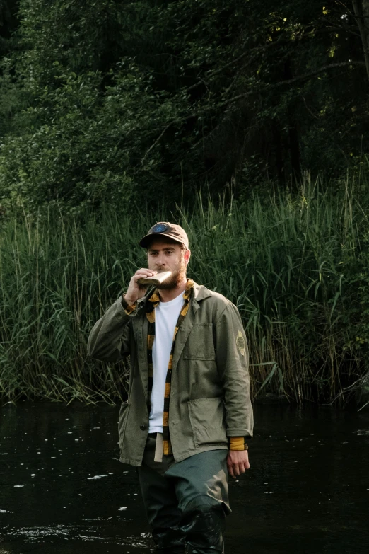 a man standing in a river with a fish in his mouth, an album cover, by Jesper Knudsen, unsplash, chris evans with a beer belly, standing in tall grass, drinking tea, streetwear